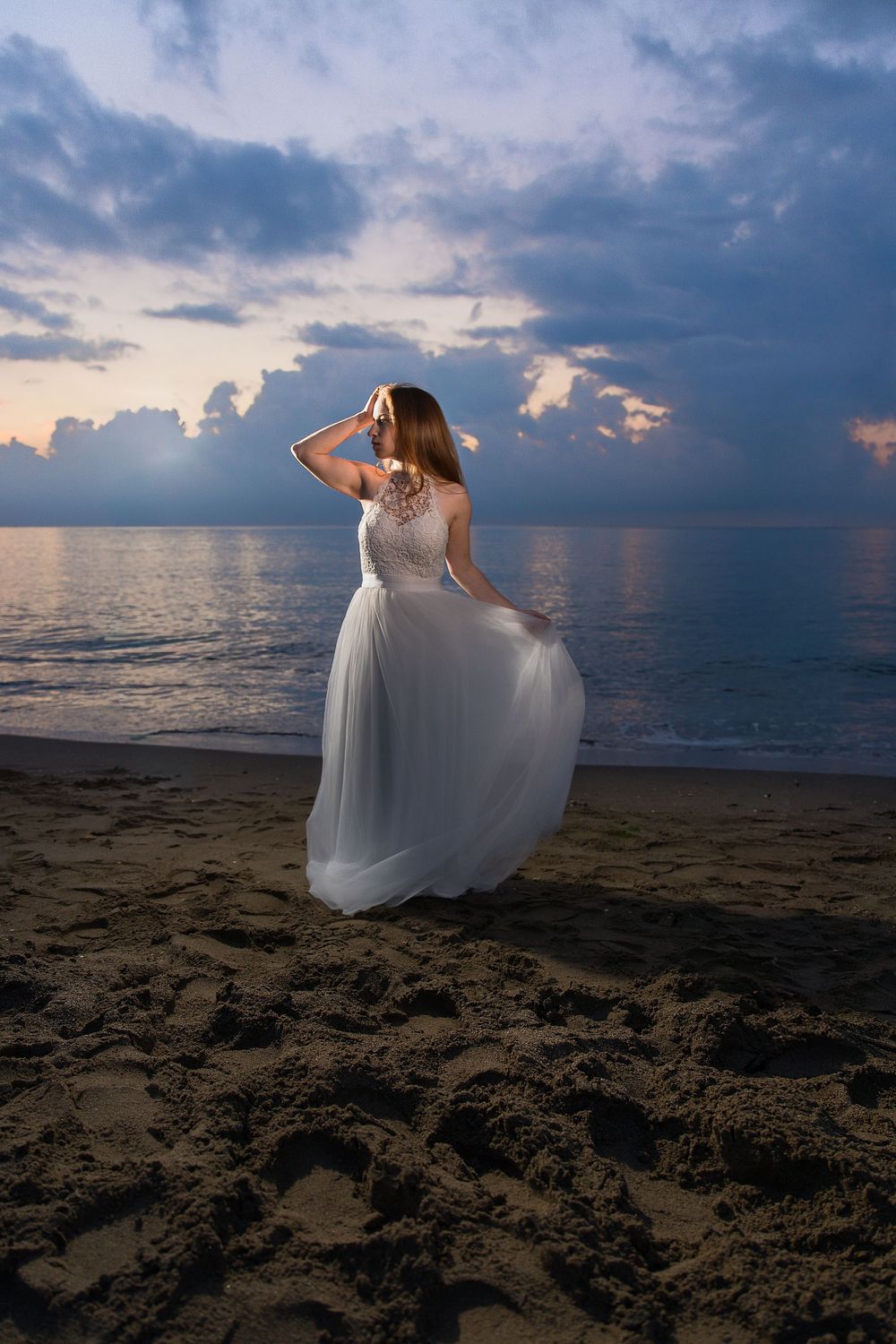 A flowing white wedding dress catches the evening light as someone poses gracefully on a beach at sunset.