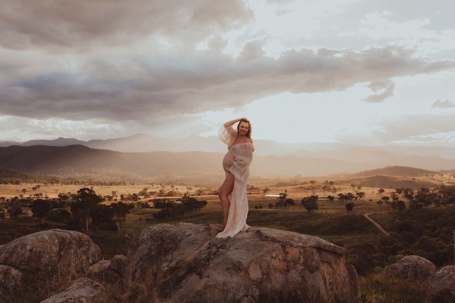 Woman in flowing white dress poses among tall grass with mountains and sunset sky.
