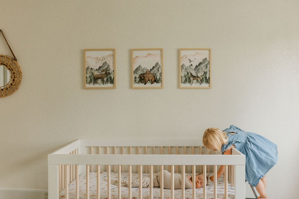 A toddler in a blue dress leans into a white crib beneath three framed mountain art prints in a neutral nursery.