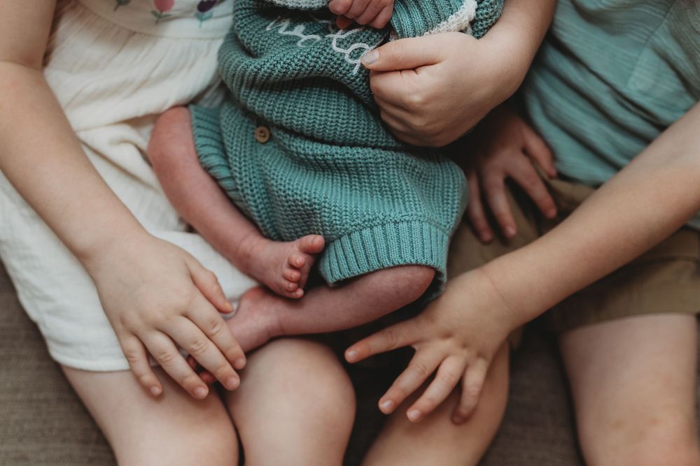 Newborn baby in green knit outfit held by two young siblings, showing tiny feet and hands close-up.