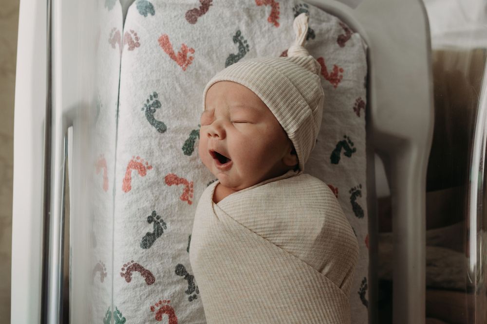 Newborn baby yawning, swaddled in a cream knit blanket, wearing a beige hat, lying in a hospital bassinet.