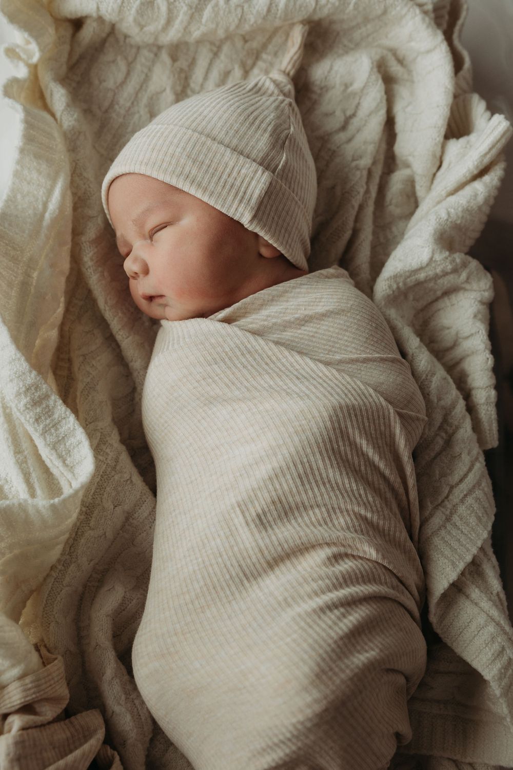 Peaceful newborn baby sleeping swaddled in cream ribbed wrap and matching knot hat on soft white blanket.