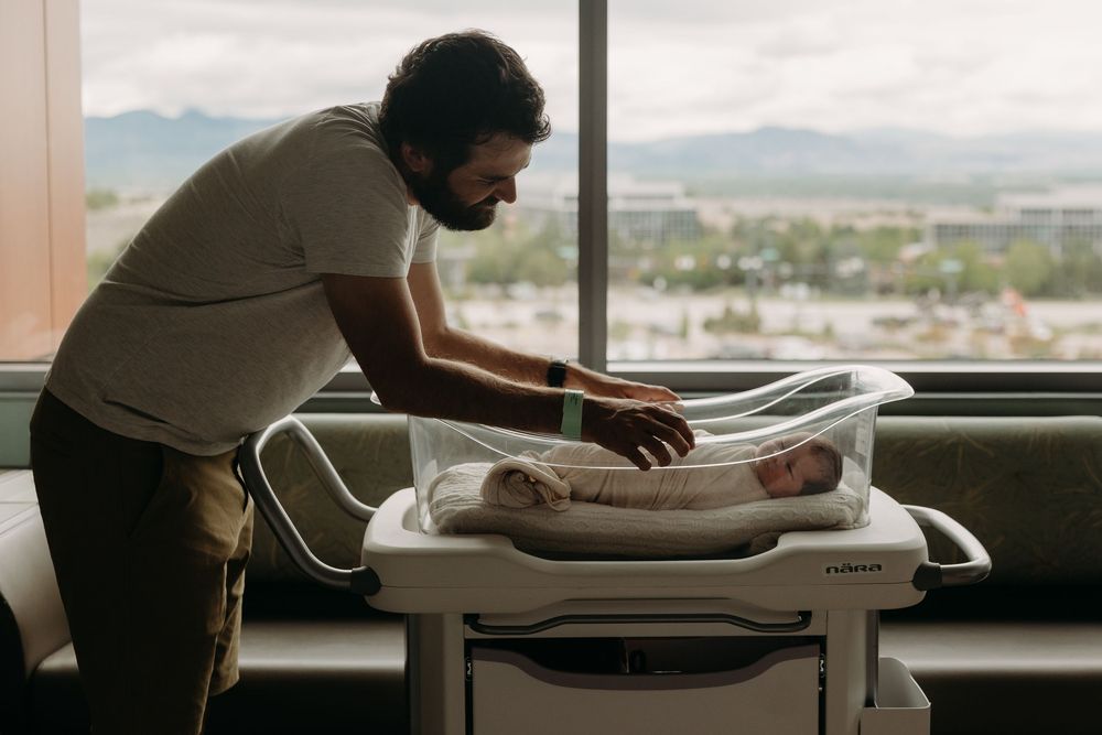Father tenderly caring for newborn baby in hospital bassinet with mountain view window backdrop.