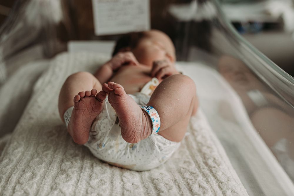 Newborn baby lying in hospital bassinet wearing diaper and ID bracelet, tiny feet visible up close.