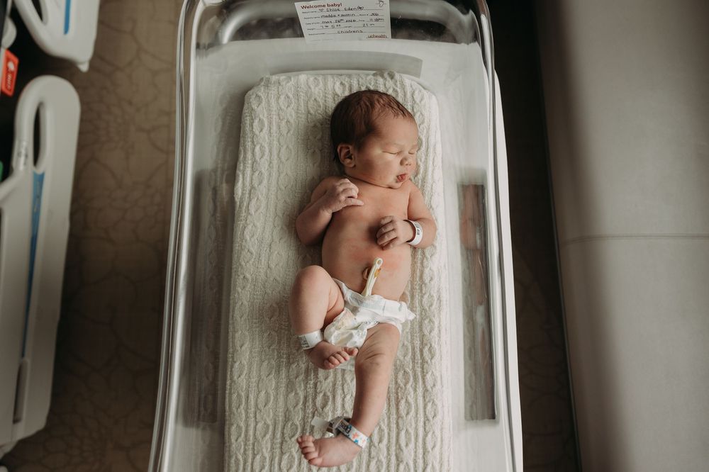 Newborn baby lying in a hospital bassinet wearing a diaper, with identification tags on wrist and ankle.