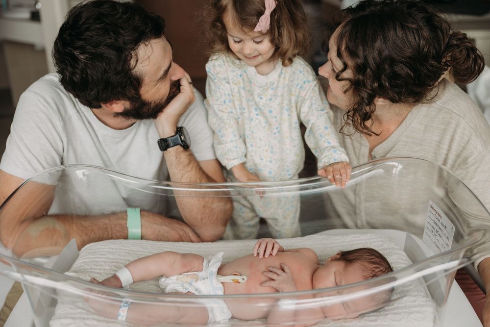Family with toddler girl admiring newborn baby lying in hospital bassinet, tender first meeting moment.