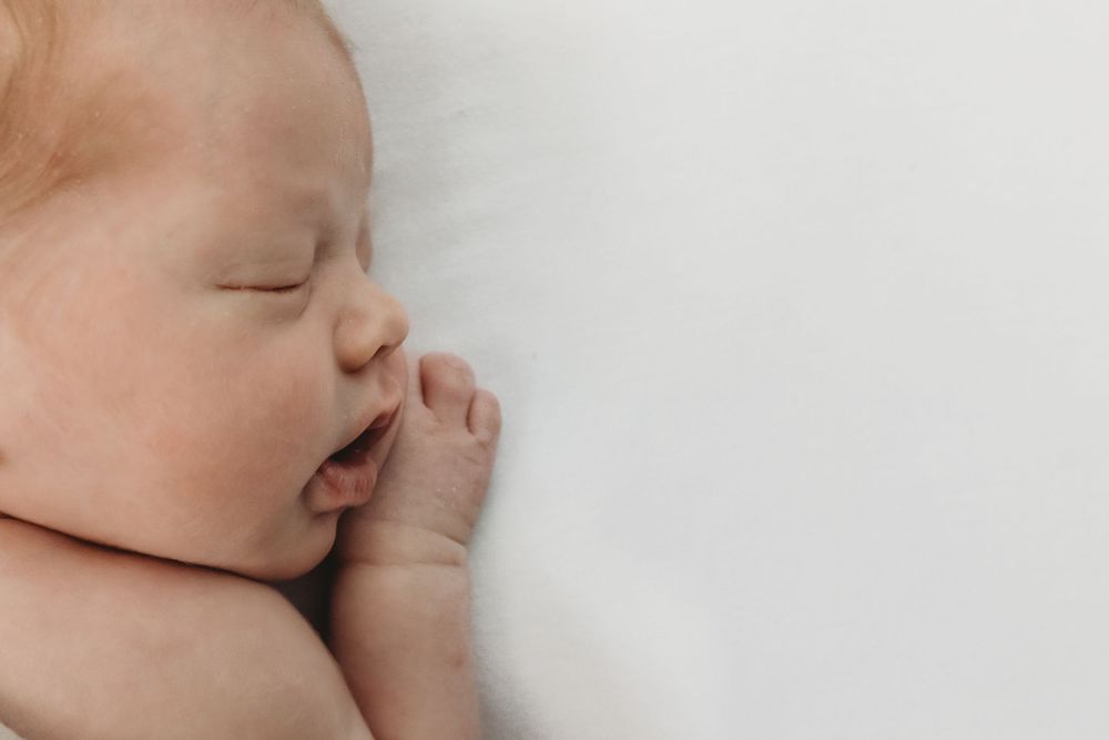 Peaceful newborn baby sleeping on white blanket with tiny hand near face in soft natural light.