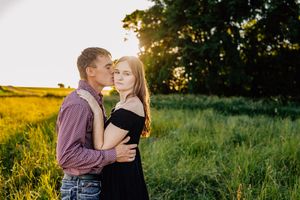 A couple embraces in a sunlit meadow during golden hour with trees and tall grass in the background.