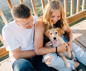 Young couple sits on wooden deck petting their smiling tan puppy on a sunny afternoon.