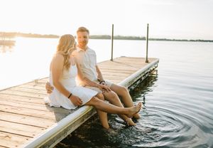 A couple sits together on a wooden dock by the water during sunset, dangling their feet over the edge.