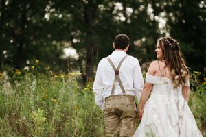 A couple in rustic wedding attire walks together through a sunlit wildflower field.