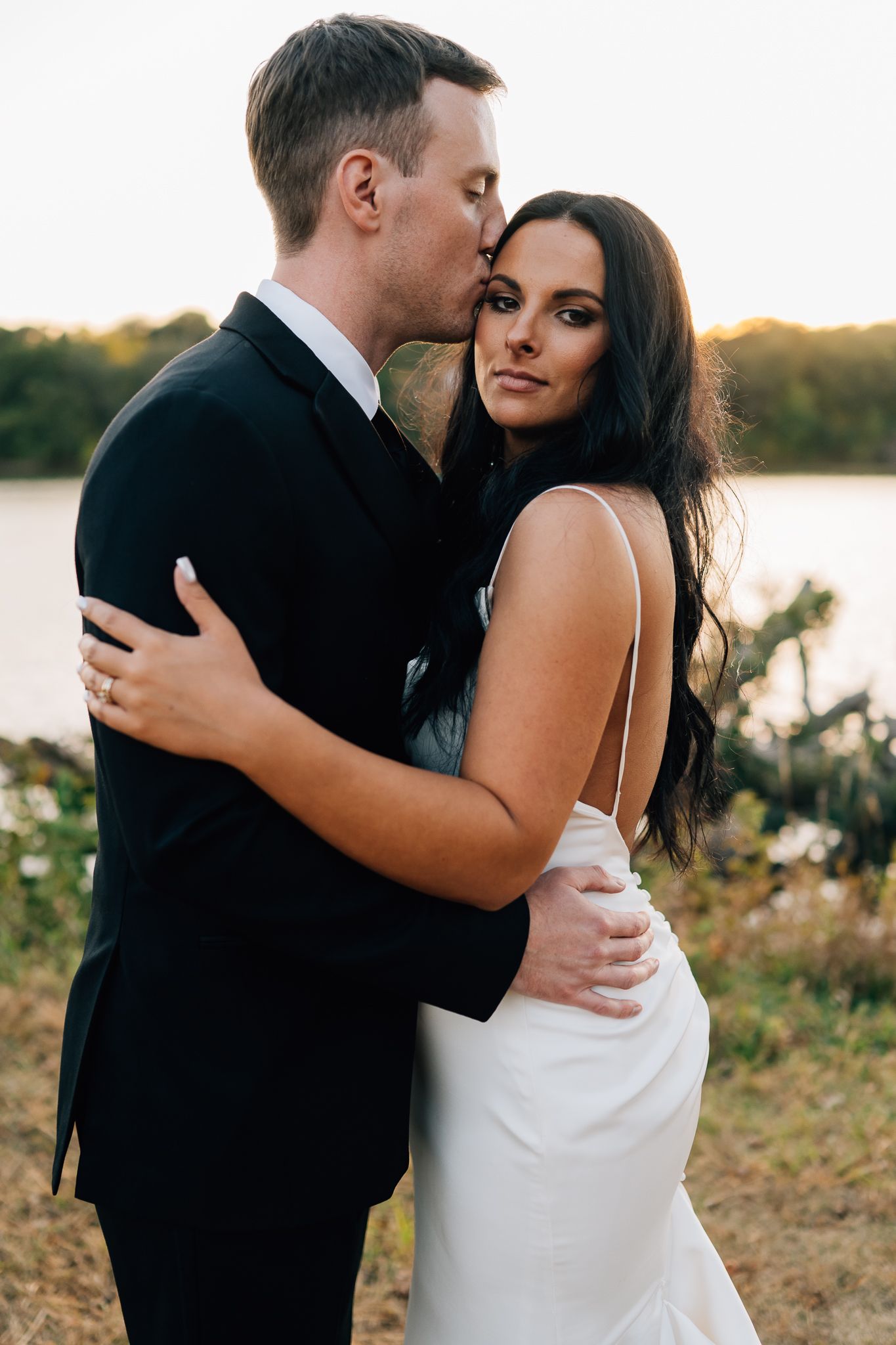 Elegant couple in wedding attire embracing during sunset photoshoot near water.