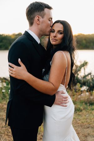Elegant couple in wedding attire embracing during sunset photoshoot near water.