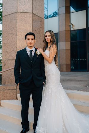 A couple in formal attire poses near a modern building entrance with stone columns.