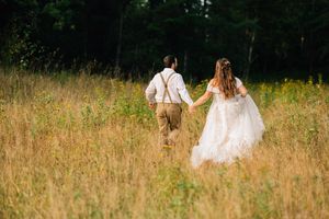 A couple in wedding attire walks hand in hand through a sunlit meadow of tall golden grass.