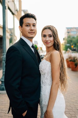 A couple in formal wedding attire pose together on a brick sidewalk during golden hour.