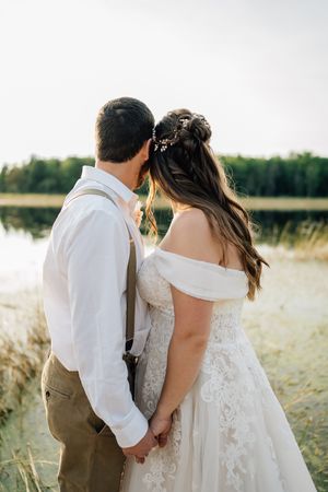 A romantic couple in wedding attire holding hands by a lake at sunset.