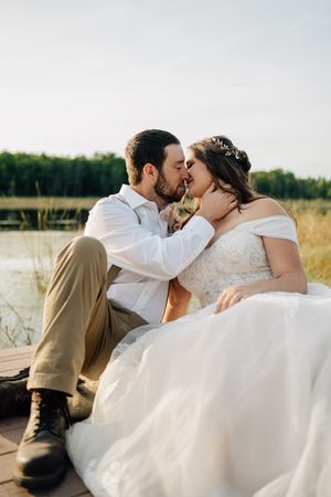 A couple shares an intimate moment sitting on a dock beside a lake in formal wedding attire.