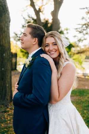 A couple in wedding attire poses together outdoors during golden hour with autumn trees in the background.