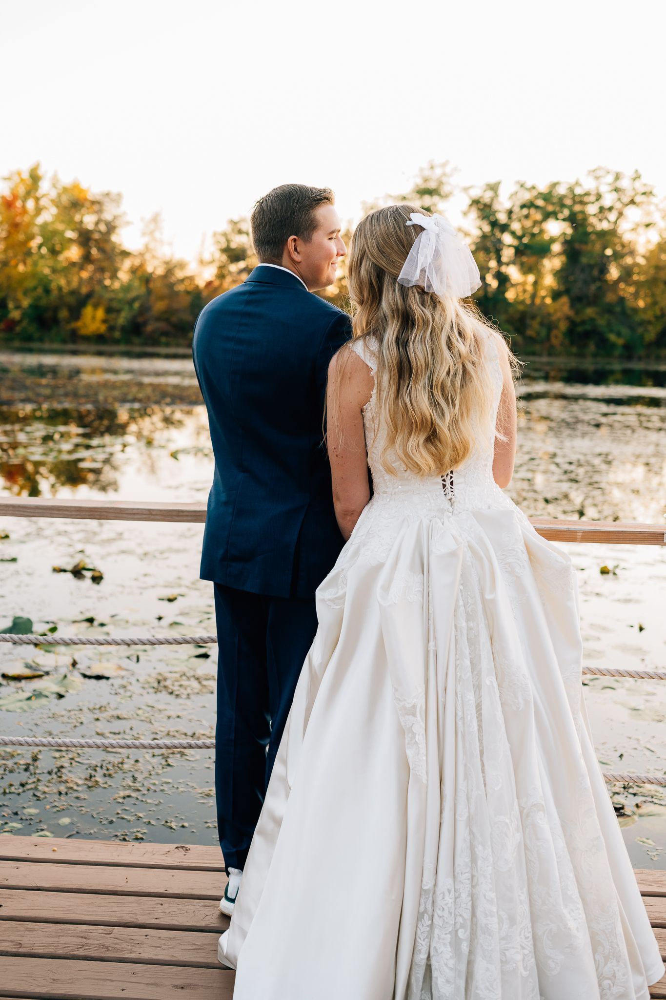 A bride in a white wedding gown and groom in navy suit stand together on a wooden deck during sunset.
