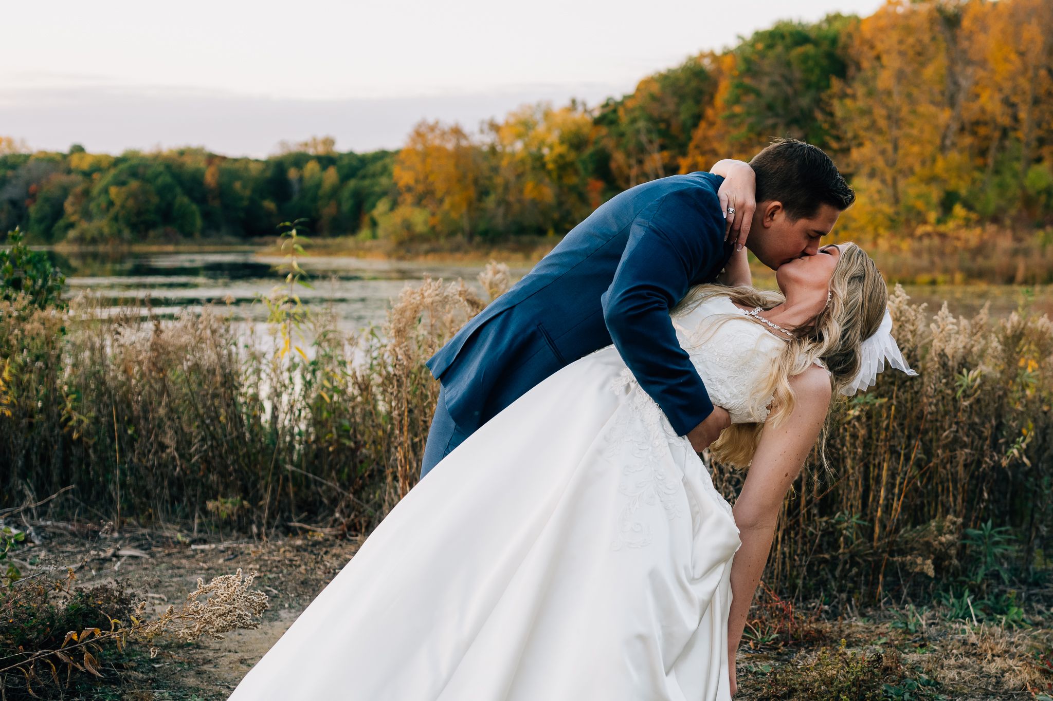A romantic dip kiss between newlyweds in formal attire by an autumn lake.