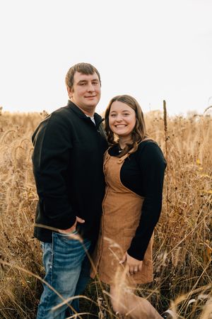 A couple in matching black sweaters stands intimately together in a golden autumn field during sunset.