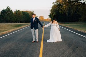 Newlyweds hold hands while standing on opposite sides of a yellow road line at sunset.