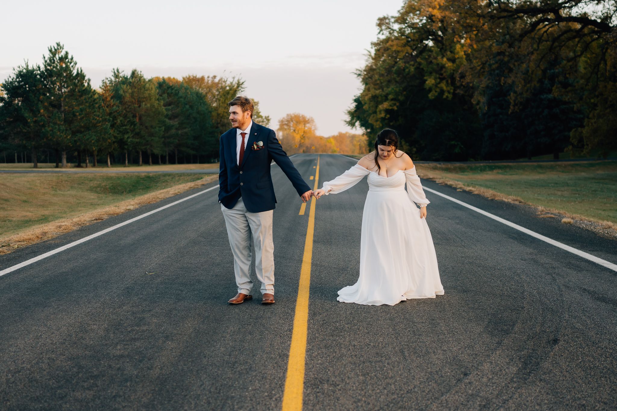 Newlyweds hold hands while standing on opposite sides of a yellow road line at sunset.