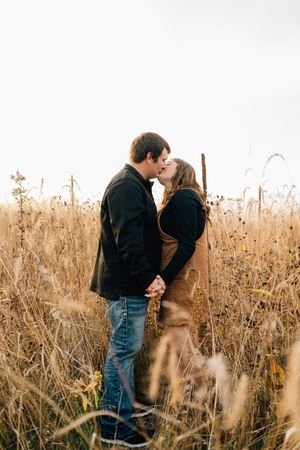 A romantic couple shares a tender kiss while standing in a field of tall autumn grass at sunset.