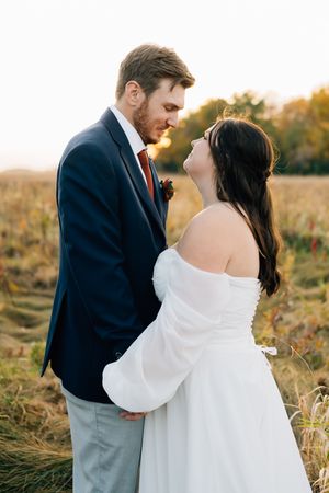 Intimate wedding portrait of couple in formal attire standing in golden field at sunset.