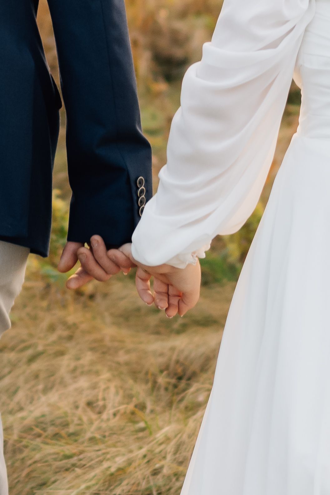 Close-up of newlywed couple holding hands, showing navy suit sleeve and white wedding dress against golden field.