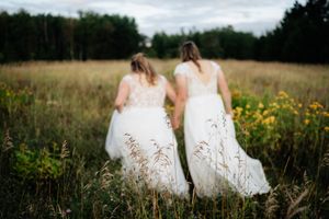 Two people in matching white dresses walk together through a wildflower field at sunset.