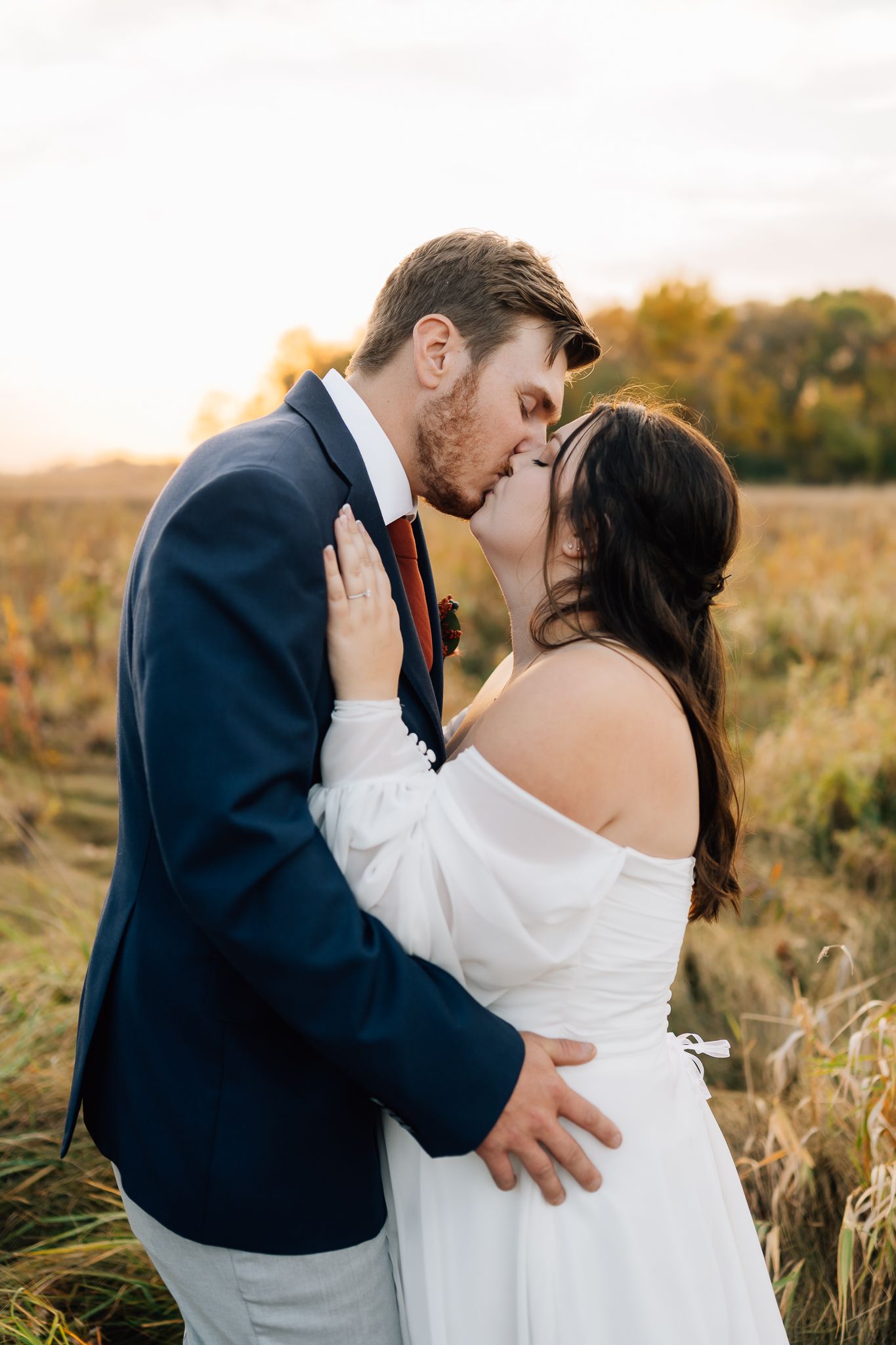A romantic couple shares a kiss in a field at golden hour, with the bride in white and groom in navy suit.