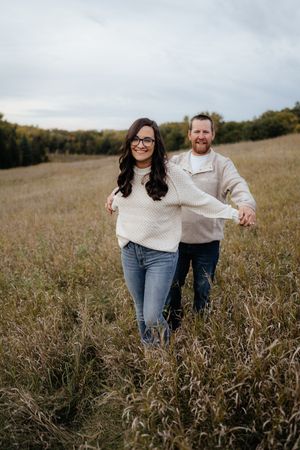 A couple wearing matching cream sweaters holds hands while walking through a rustic field.