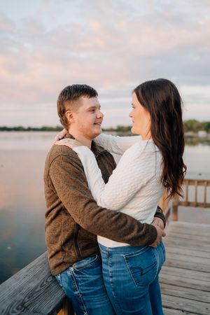 A romantic couple in sweaters embrace on a wooden dock by a lake during sunset.