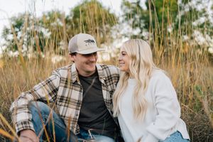 Happy couple in casual flannel shirts sitting together in tall grass during sunset.