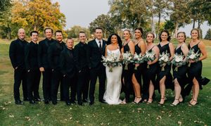 A wedding party in black and navy attire poses together outdoors during autumn.