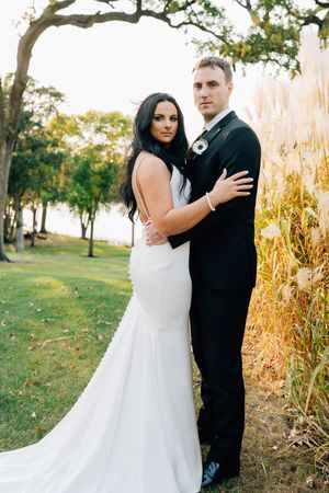 A bride in a white mermaid dress and groom in black suit pose together outdoors near tall ornamental grasses.