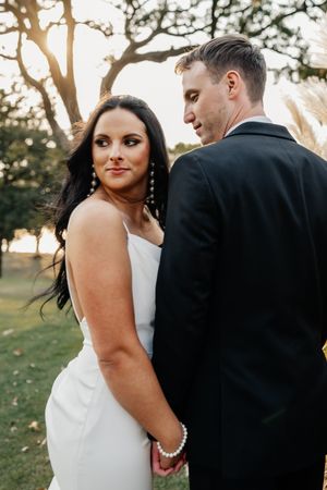 Elegant bride in sleek white dress looks over shoulder while holding groom's hand at outdoor venue.