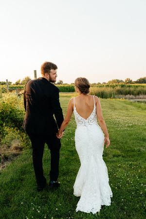 A couple walks hand in hand through a grassy field with wildflowers at golden hour.