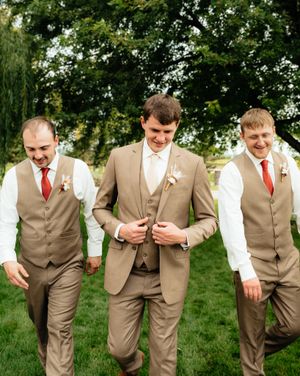 Groomsmen in tan suits and vests share a candid moment on a lawn with trees in the background.