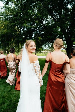 A bride in a lace gown with bridesmaids in burgundy dresses walking on grass.