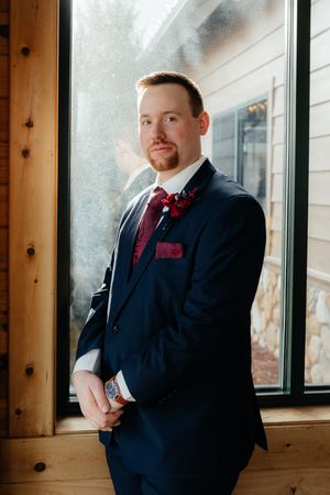 A well-dressed person in a navy suit with burgundy bow tie stands confidently by a sunny window in a rustic room.