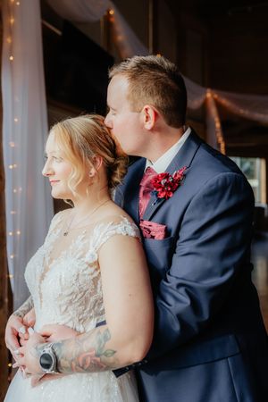 Sweet moment between newlyweds as groom kisses bride's head, both showing off tattoos and formal attire.