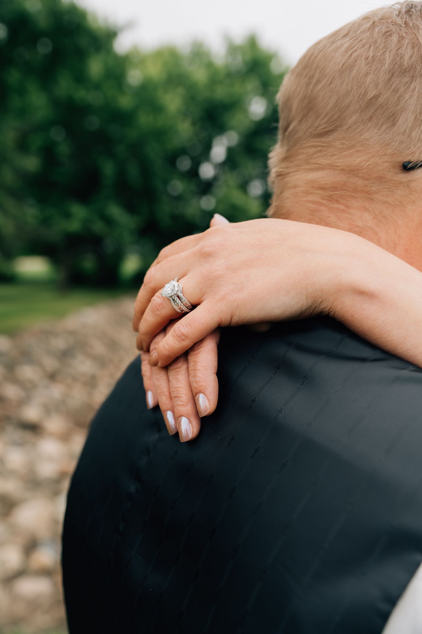 Close-up view of an engagement ring and hands embracing against a black jacket outdoors.