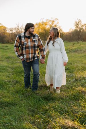 A couple walks through tall grass at sunset, one in a plaid shirt and the other in a white dress.