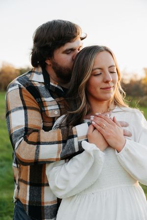 A romantic couple embraces in a field during golden hour, with one wearing a plaid shirt and the other in white.