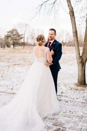 A couple shares an intimate winter wedding moment on a snowy brick pathway surrounded by bare trees.