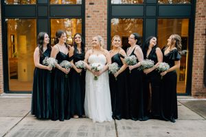 Bridal party in navy blue velvet dresses poses with bride in white gown outside brick building.