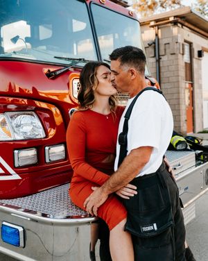 Couple poses against red fire truck in romantic engagement session.